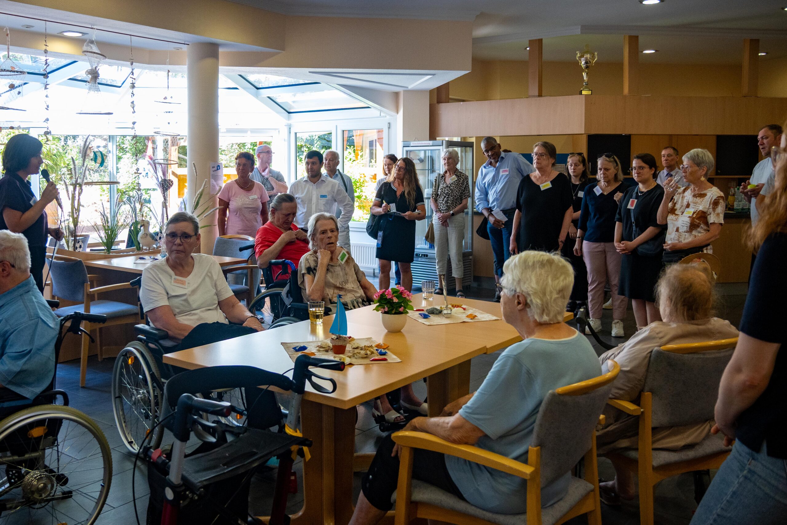 Eine Rednerin spricht vor Senioren und weiteren Gästen der Veranstaltung bei einer Veranstaltung in einem hellen Café; die Teilnehmer sitzen in Rollstühlen oder auf Stühlen um einen Tisch mit Erfrischungen herum.
