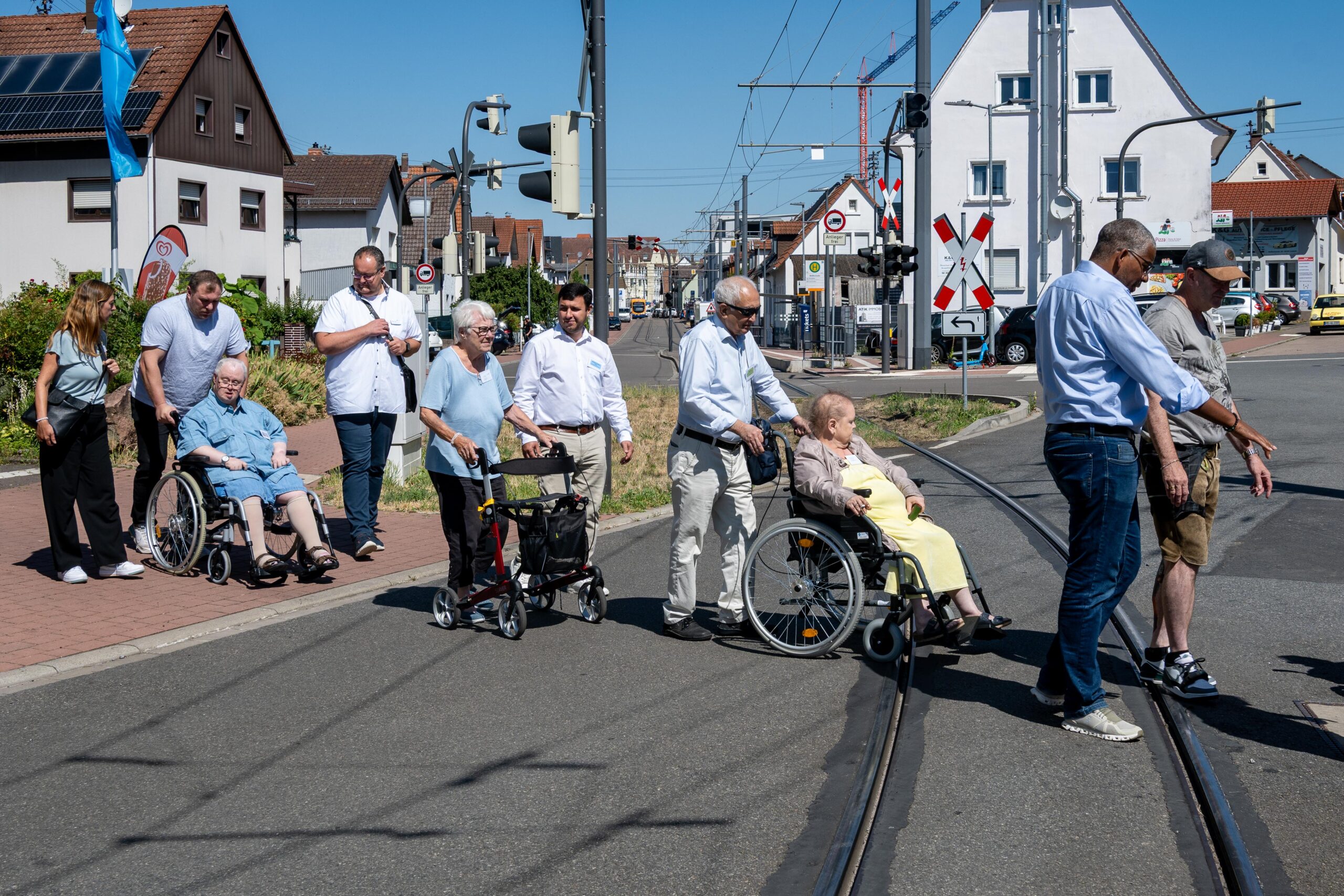 Im Bild dargestellt ist der Spaziergang zur Barrierefreiheit in Leimen. Eine Gruppe von Menschen, zwei davon im Rollstuhl und eine Person mit Rollator. Sie überqueren eine Strasse mit Strassenbahnschienen.