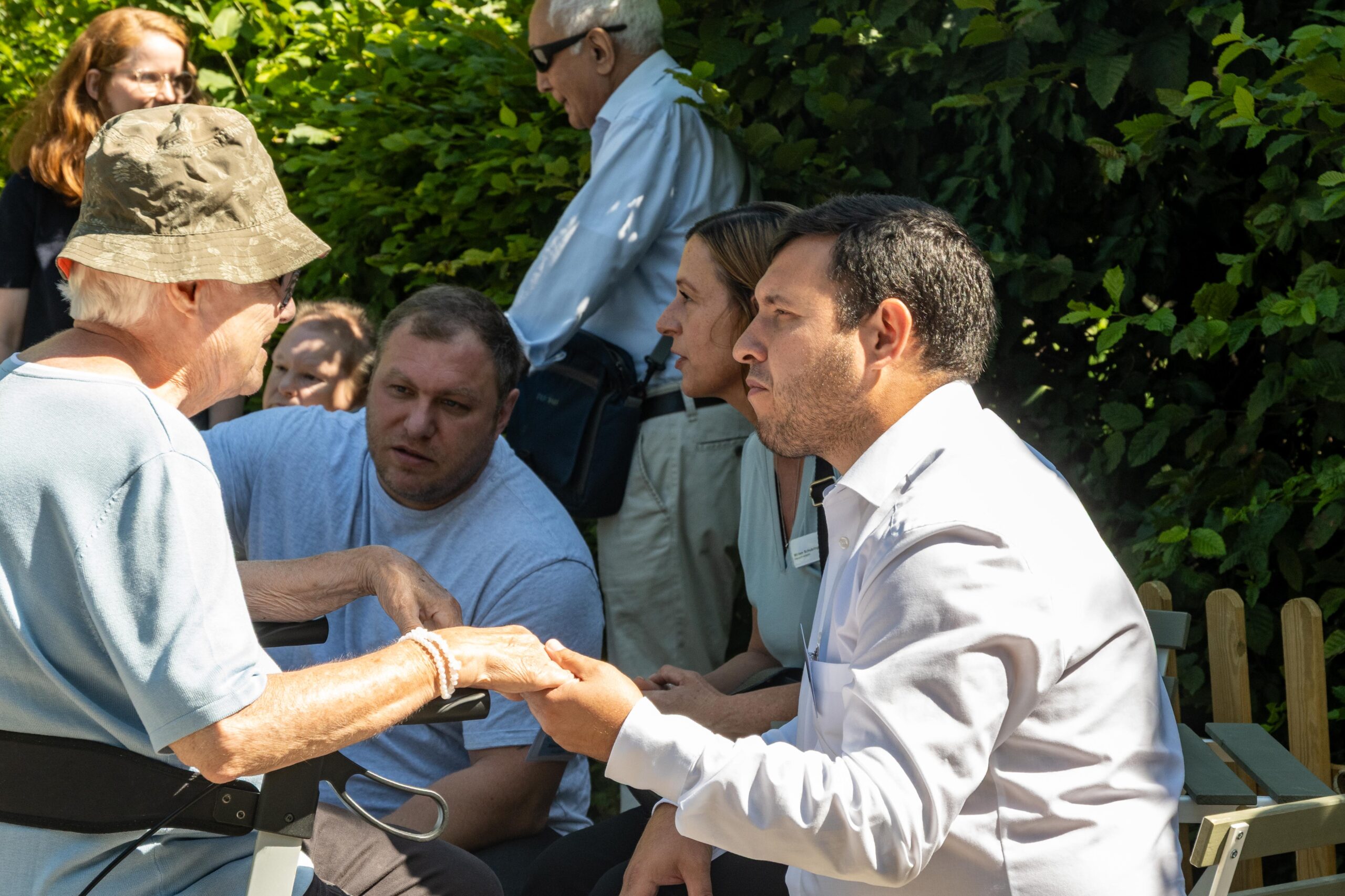 Das Bild zeigt eine Gruppe von Menschen, die im Schatten einer Hecke sitzt. Im Vordergrund hält ein jüngerer Mann die Hände einer älteren Dame, die auf ihrem Rollator sitzt.