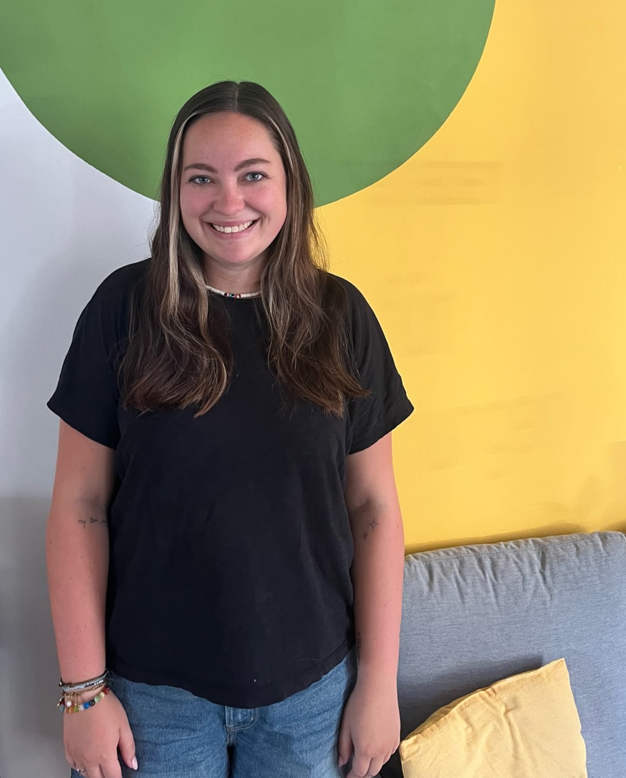 Alissa Stein_Team_Quartiersmanagement Leimen Eine junge Frau in schwarzem T-Shirt und Jeans steht vor einer gelb-grünen Wand.