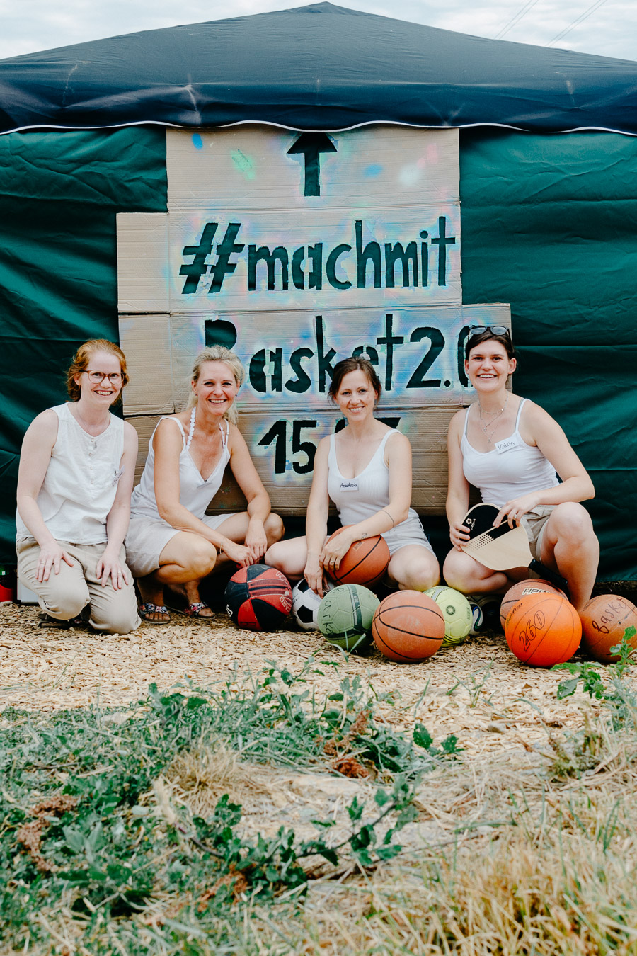 2023-07-15-Jugend ist gefragt_004-kl Vier Frauen sitzen vor einem selbst gestalteten Schild mit der Aufschrift „#machmit – …“. Vor ihnen liegen mehrere Bälle, darunter Basket-, Fuß- und Handbälle, sowie ein Schläger. Die Frauen tragen helle Kleidung und lächeln in die Kamera. Die Aufnahme entstand im Rahmen einer Mitmach- oder Sportveranstaltung unter einem Pavillon.
