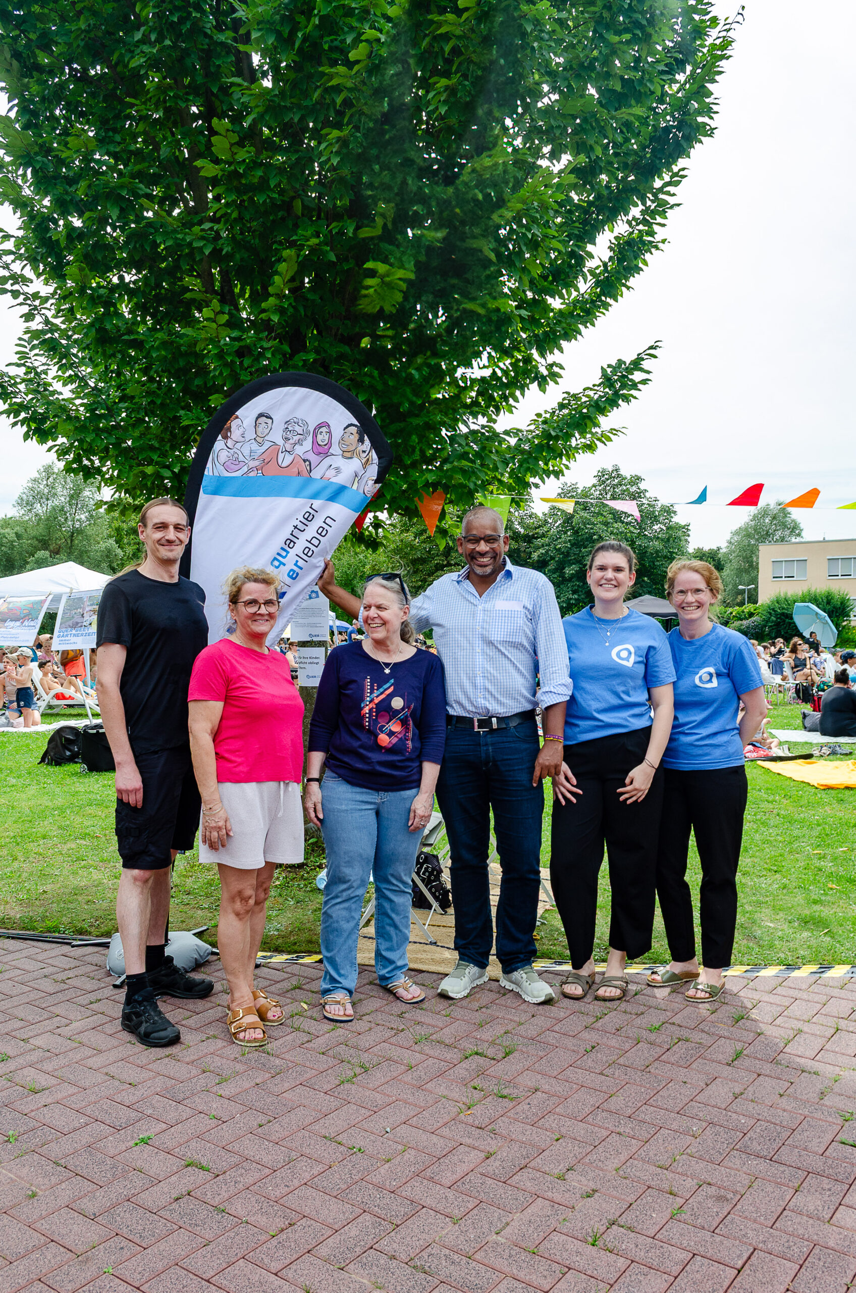 2025-07-19-Kino im Freibad_062-gr Sechs Personen stehen vor einer Beachflag mit der Aufschrift Quartier erleben. Im Hintergrund findet eine Open-Air Kinoveranstaltung statt.