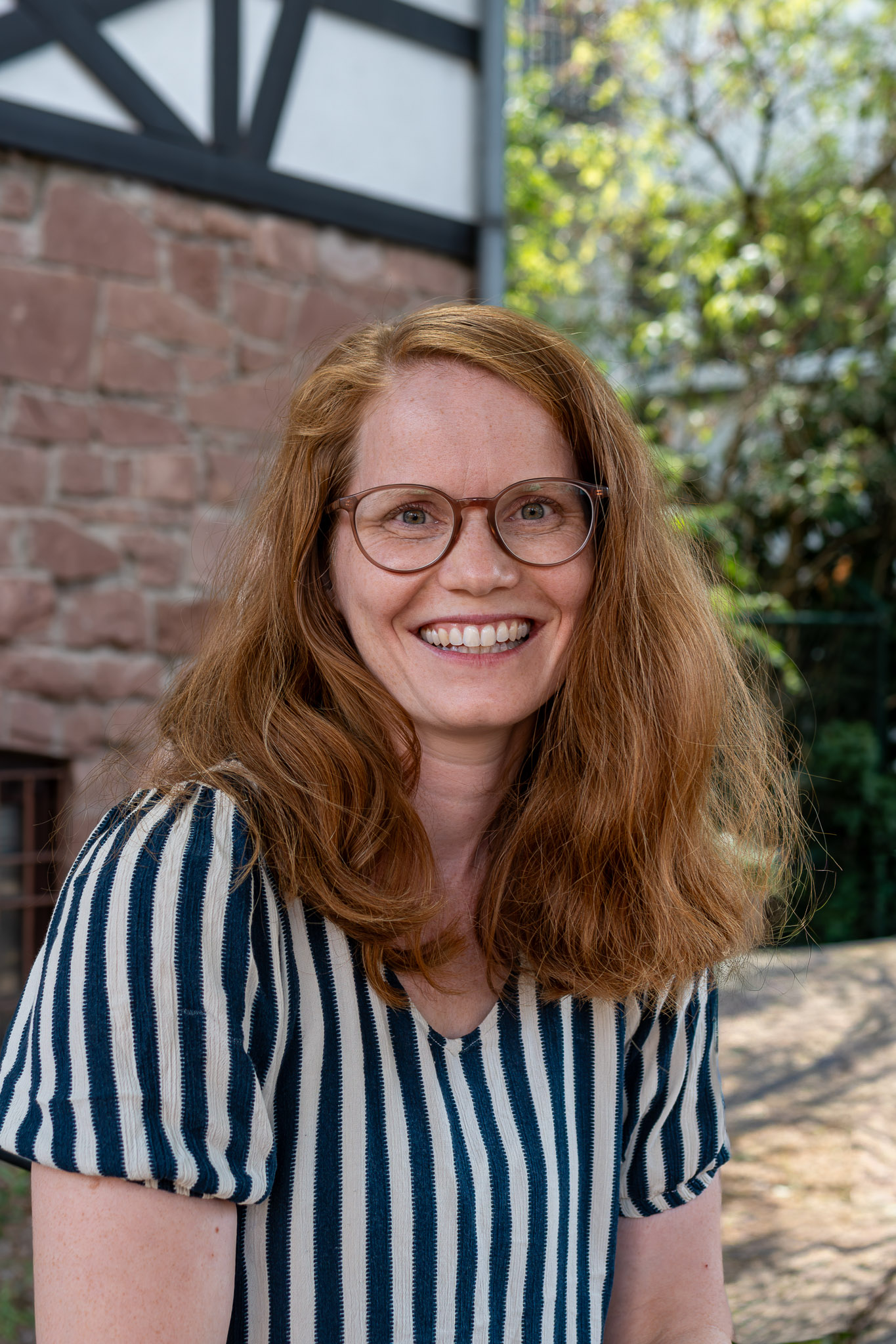 Das Foto zeigt eine Frau mit offenen roten Haaren. Sie trägt eine Brille und eine blau-weiß gestreifte Bluse.