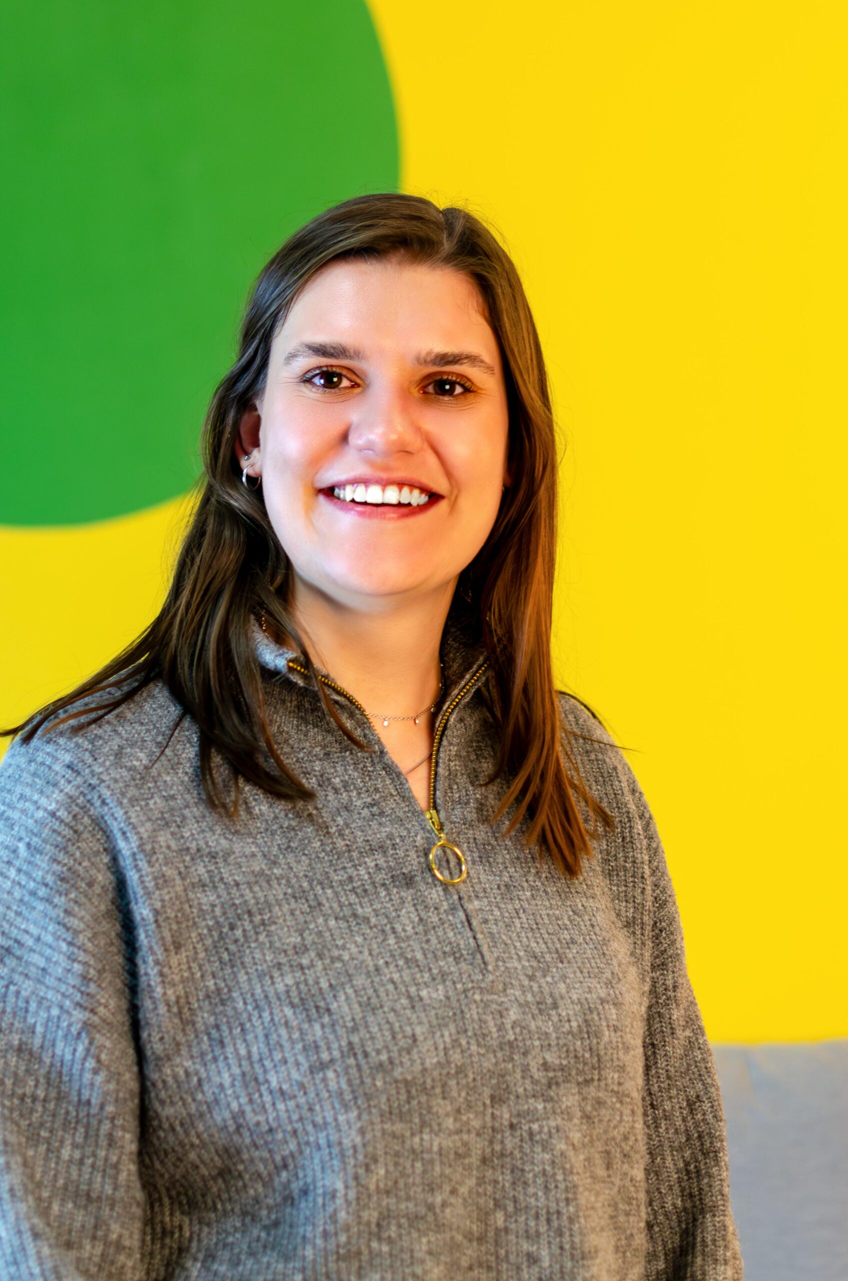 QUER-Team_Katrin_003-kl Das Bild zeigt eine junge Frau mit brauen Haaren. Sie lächelt und trägt einen grauen Pullover. Die Wand im Hintergrund ist gelb und grün.